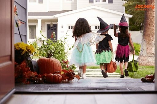  Trois enfants déguisés pour Halloween, l'un en fée et les deux autres en sorcières, marchent sur le porche d'une maison décorée avec des citrouilles et des feuillages d'automne.