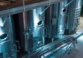 Vue plongeante sur de grandes cuves industrielles en acier inoxydable au sein d'une installation de production moderne, avec une pompe industrielle à engrenages visible au premier plan, des canalisations et un éclairage d'ambiance bleuté.