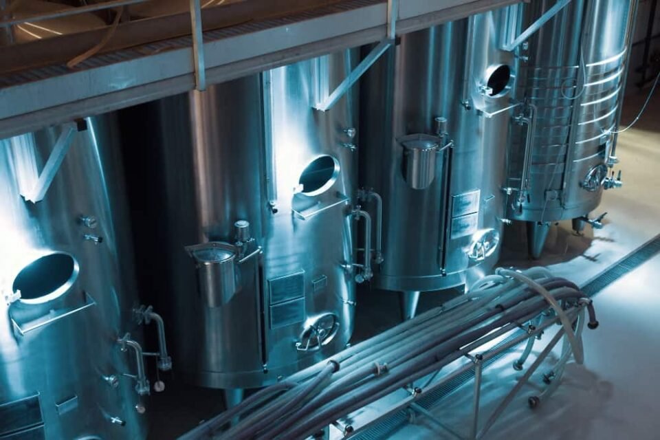 Vue plongeante sur de grandes cuves industrielles en acier inoxydable au sein d'une installation de production moderne, avec une pompe industrielle à engrenages visible au premier plan, des canalisations et un éclairage d'ambiance bleuté.