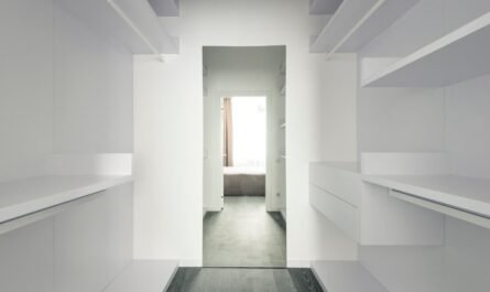 Corridor avec étagères blanches, sol gris, ouverture vers une pièce lumineuse avec lit