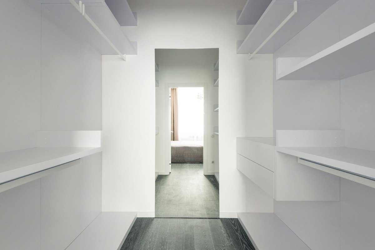 Corridor avec étagères blanches, sol gris, ouverture vers une pièce lumineuse avec lit