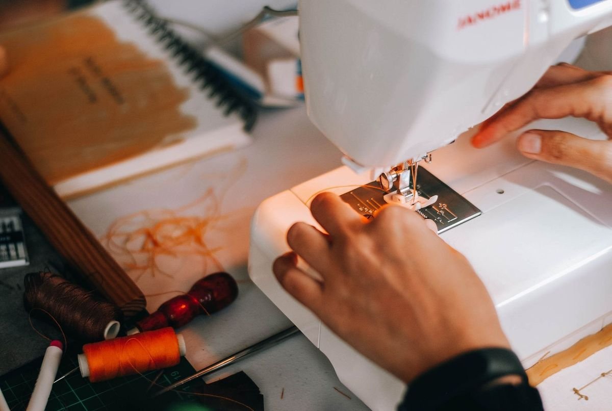 Mains guidant un tissu sous la tête d'une machine à coudre, fil orange et ciseaux à proximité